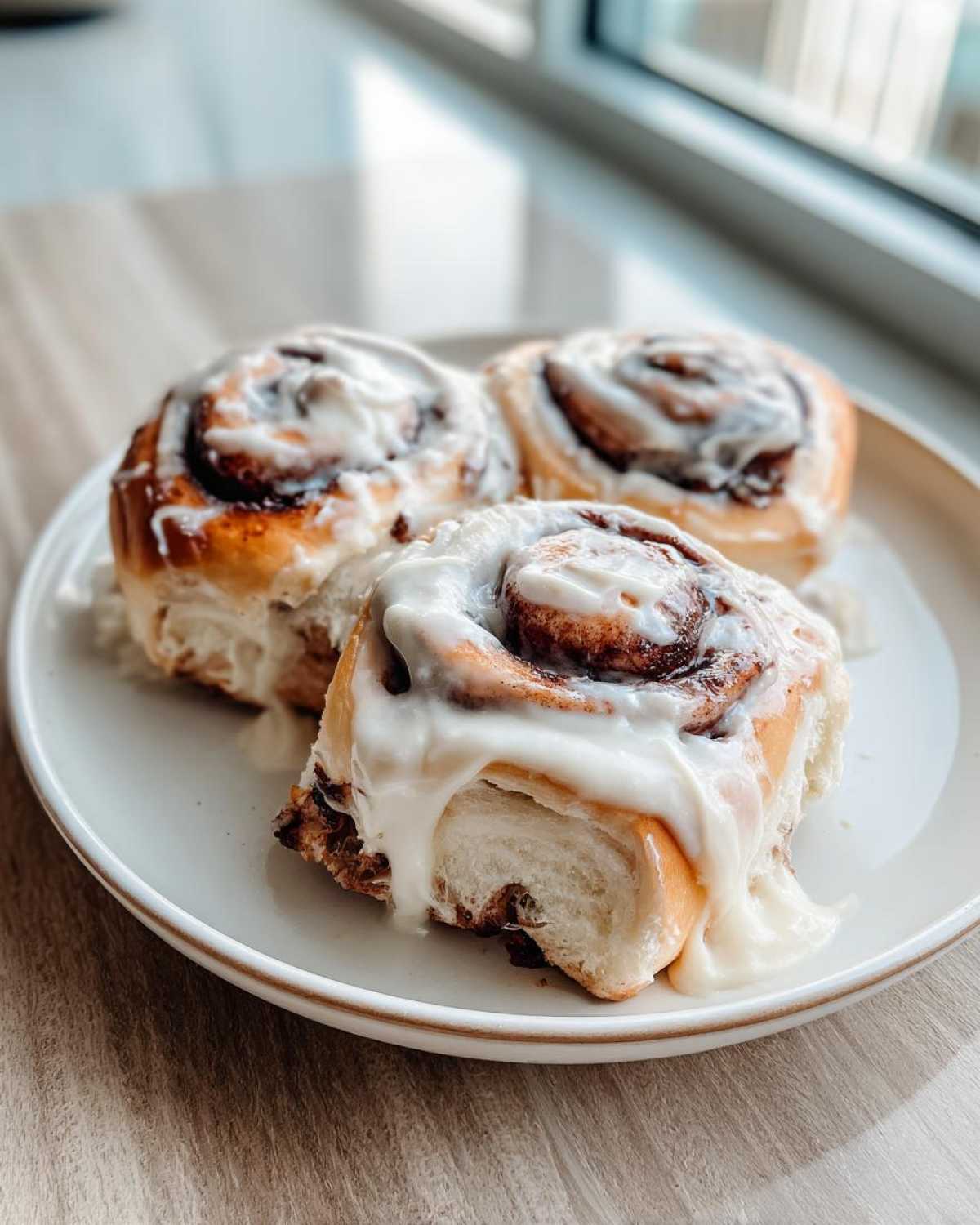 Three freshly baked sourdough discard cinnamon rolls generously topped with white cream cheese frosting, sitting on a light plate.