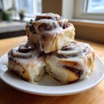Three fluffy sourdough discard cinnamon rolls stacked on a white plate, generously covered in white cream cheese icing.