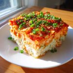 A square slice of spicy salmon sushi bake showing layers of rice and salmon, topped with green onions and sesame seeds.
