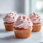 Close-up of one perfect strawberry milkshake cupcakes topped with swirled pink frosting, with two others blurred in the background.