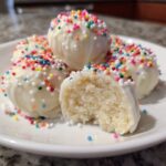 A close-up of several white chocolate coated sugar cookie truffles, one bitten open to show the dense interior, topped with colorful sprinkles.