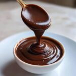 A golden spoon pours thick, glossy homemade chocolate syrup into a small white bowl, creating ripples.