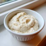 A small white ramekin filled with thick, creamy white pizza sauce, seasoned with pepper.