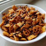 A close-up of a white bowl filled with homemade toffee chex mix recipe pieces and mini pretzels, drizzled with chocolate.