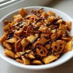 A close-up of a white bowl filled with homemade toffee chex mix recipe pieces and mini pretzels, drizzled with chocolate.