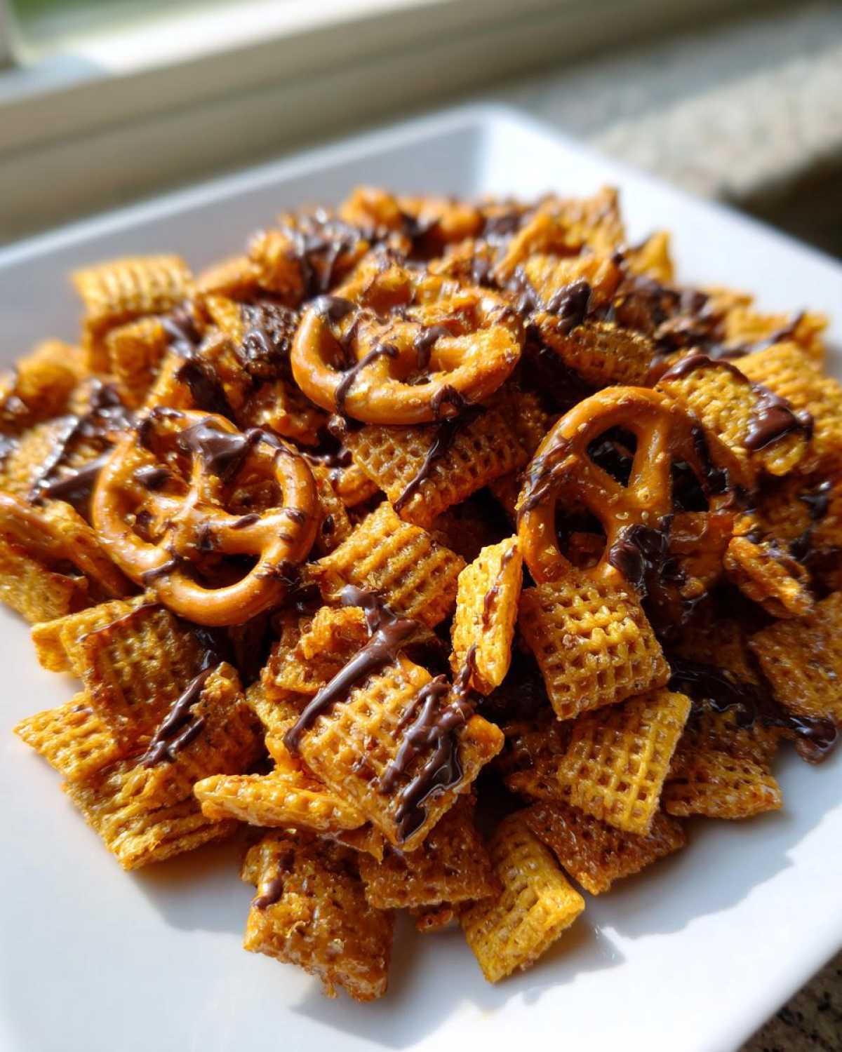 A close-up of a white plate piled high with golden toffee chex mix recipe pieces and pretzels drizzled with dark chocolate.