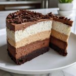 A decadent slice of triple chocolate mousse cake showing four distinct layers of chocolate mousse and a dark brownie base, topped with chocolate shavings.