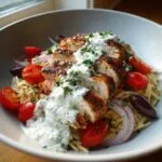 Close-up of ultimate chicken tzatziki bowls featuring sliced seasoned chicken over orzo pasta, topped with tzatziki sauce, tomatoes, and olives.