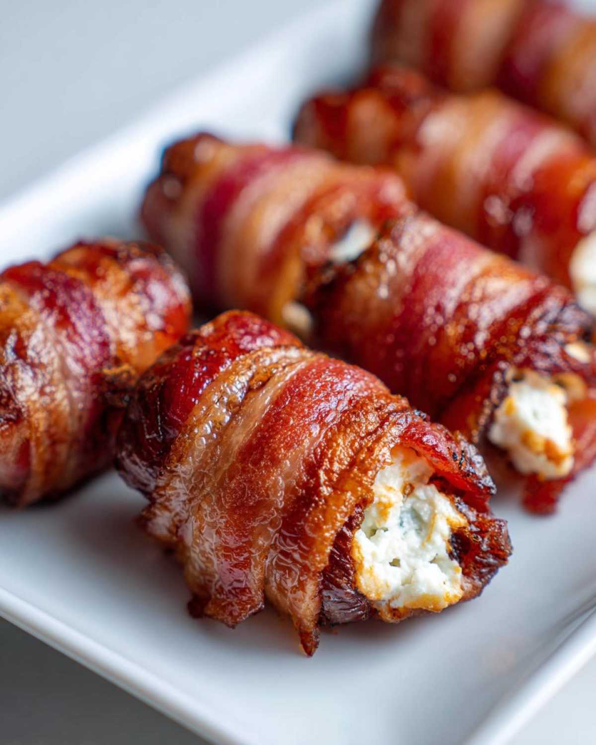 Close-up of perfectly cooked bacon wrapped dates with goat cheese spilling out, served on a white platter.