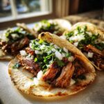 Close-up of several Tacos árabes served on grilled corn tortillas, topped generously with shredded meat, diced white onion, and fresh cilantro.