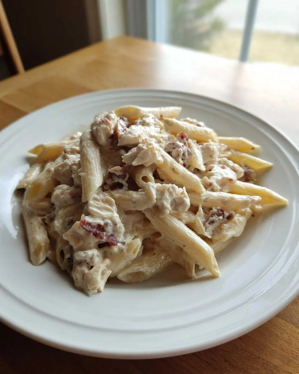 A close-up of a white plate filled with Creamy Chicken Bacon Ranch Pasta featuring penne, chicken chunks, and bacon bits.