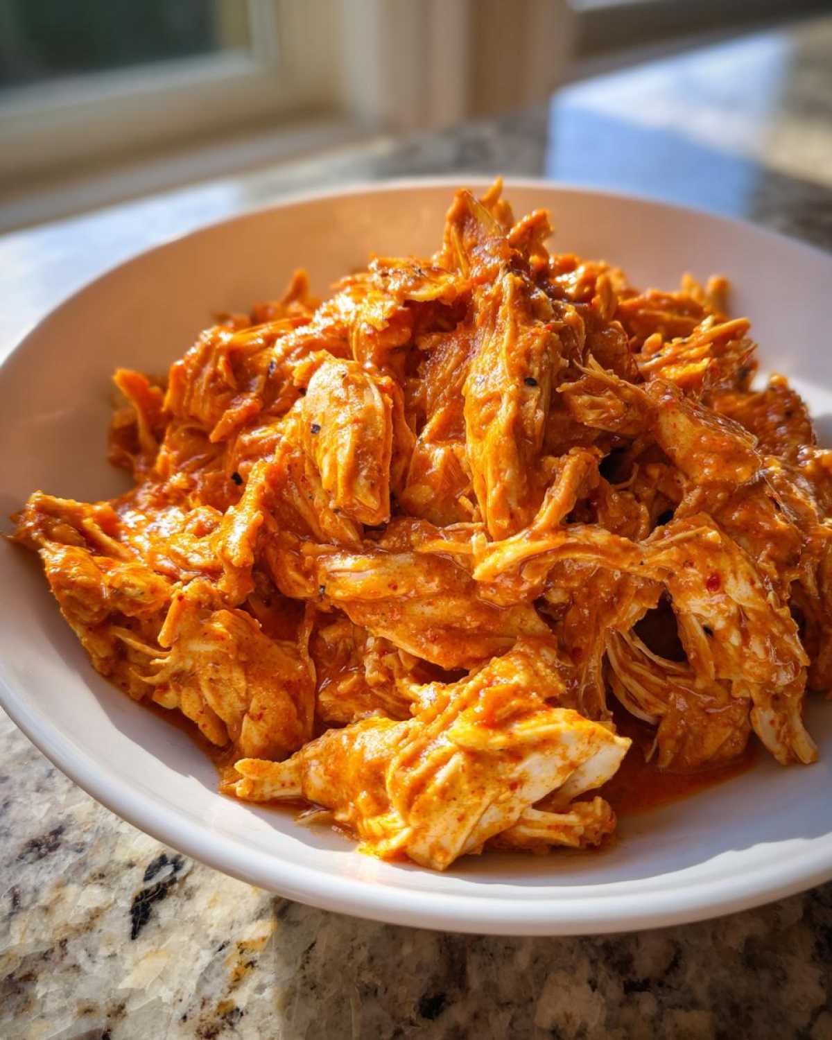 A close-up of shredded chicken coated in a rich, orange sauce, representing creamy crockpot cajun butter chicken, served in a white bowl.