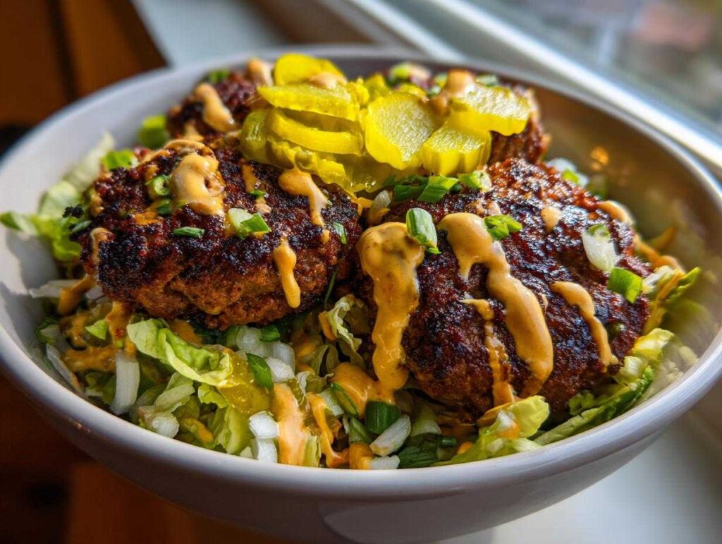 Close-up of a smash burger bowl featuring three seasoned patties over lettuce, drizzled with sauce and topped with pickles.