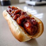 Close-up of a juicy hot dog in a bun topped with a thick, glossy, spicy red sauce for the firecracker hot dogs recipe.