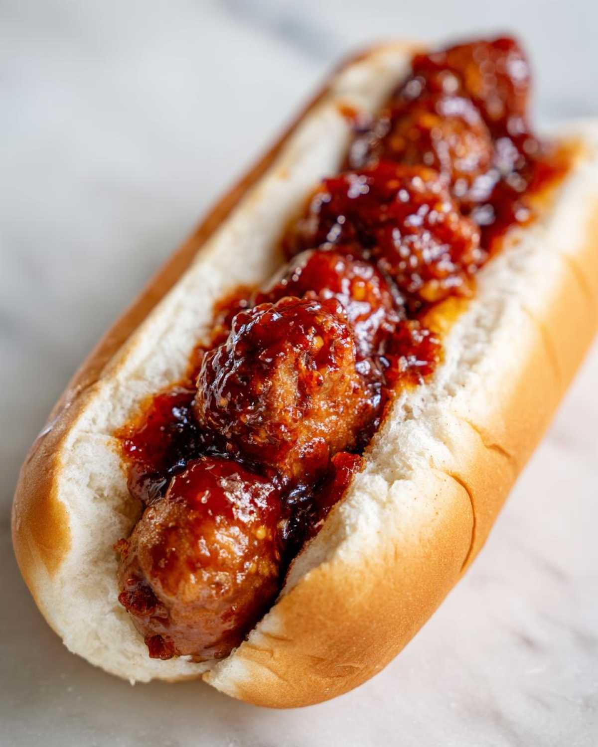 A close-up of a soft hot dog bun filled with meatballs smothered in a thick, glossy, spicy red sauce for the firecracker hot dogs recipe.