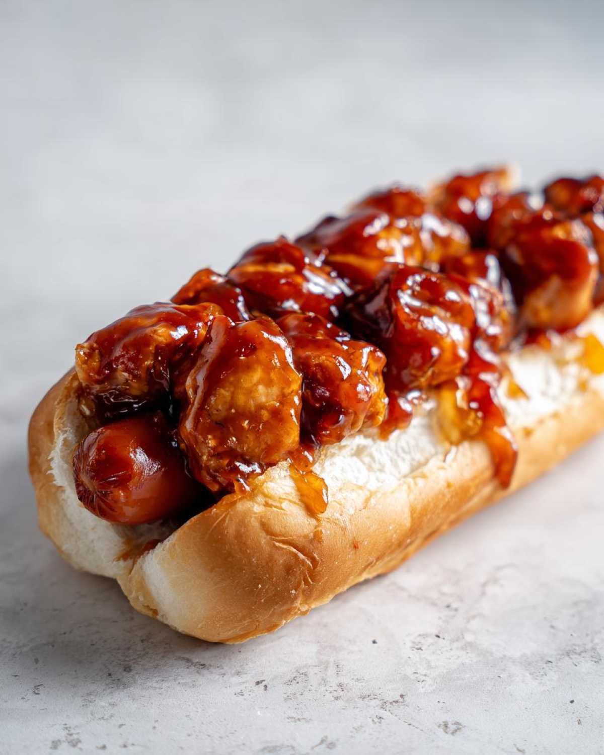 A close-up of a hot dog bun filled with a sausage, topped generously with saucy, spicy chicken pieces for the firecracker hot dogs recipe.
