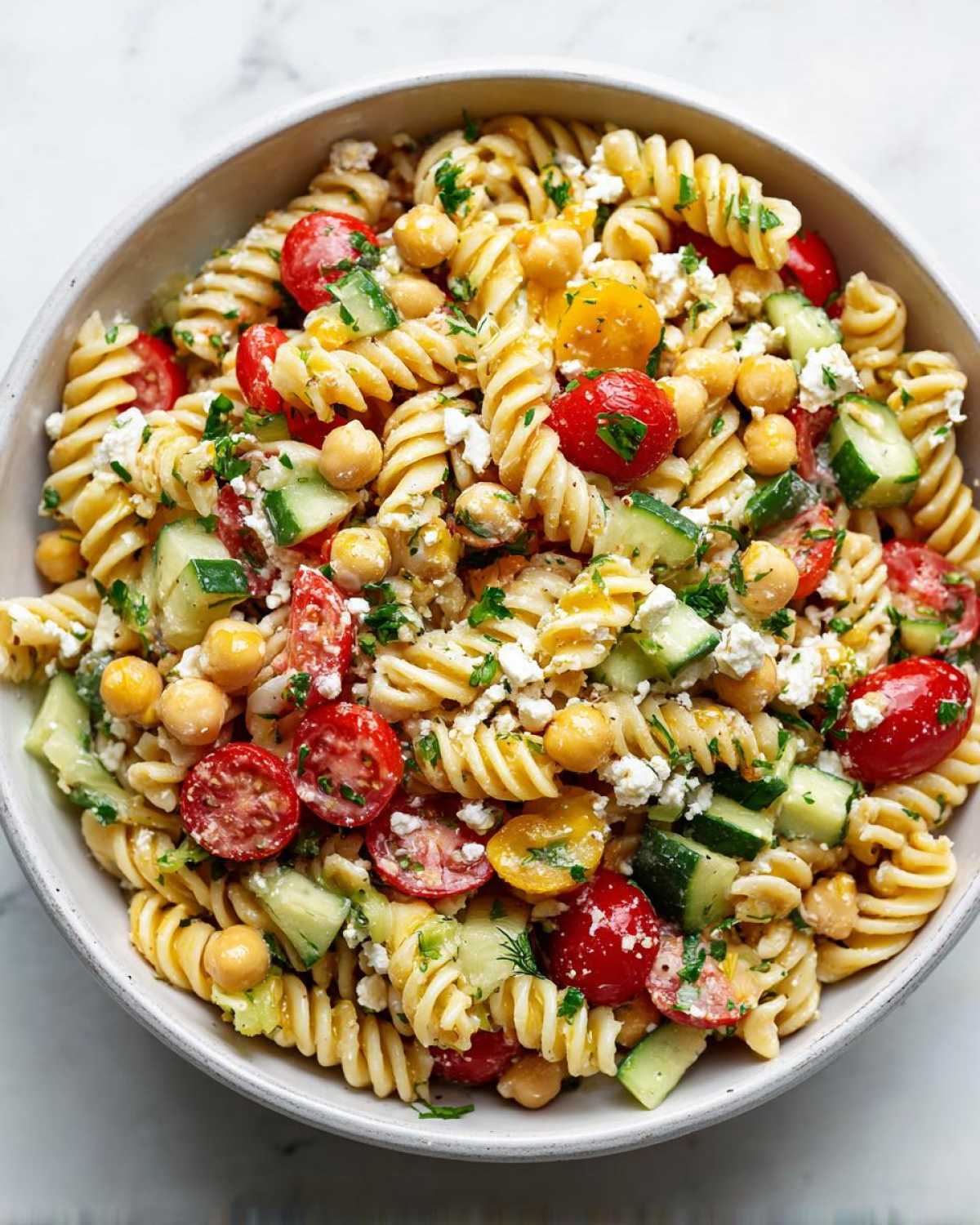 Close-up of a bowl filled with Greek pasta salad with chickpeas, featuring rotini pasta, cherry tomatoes, cucumber, and feta cheese.