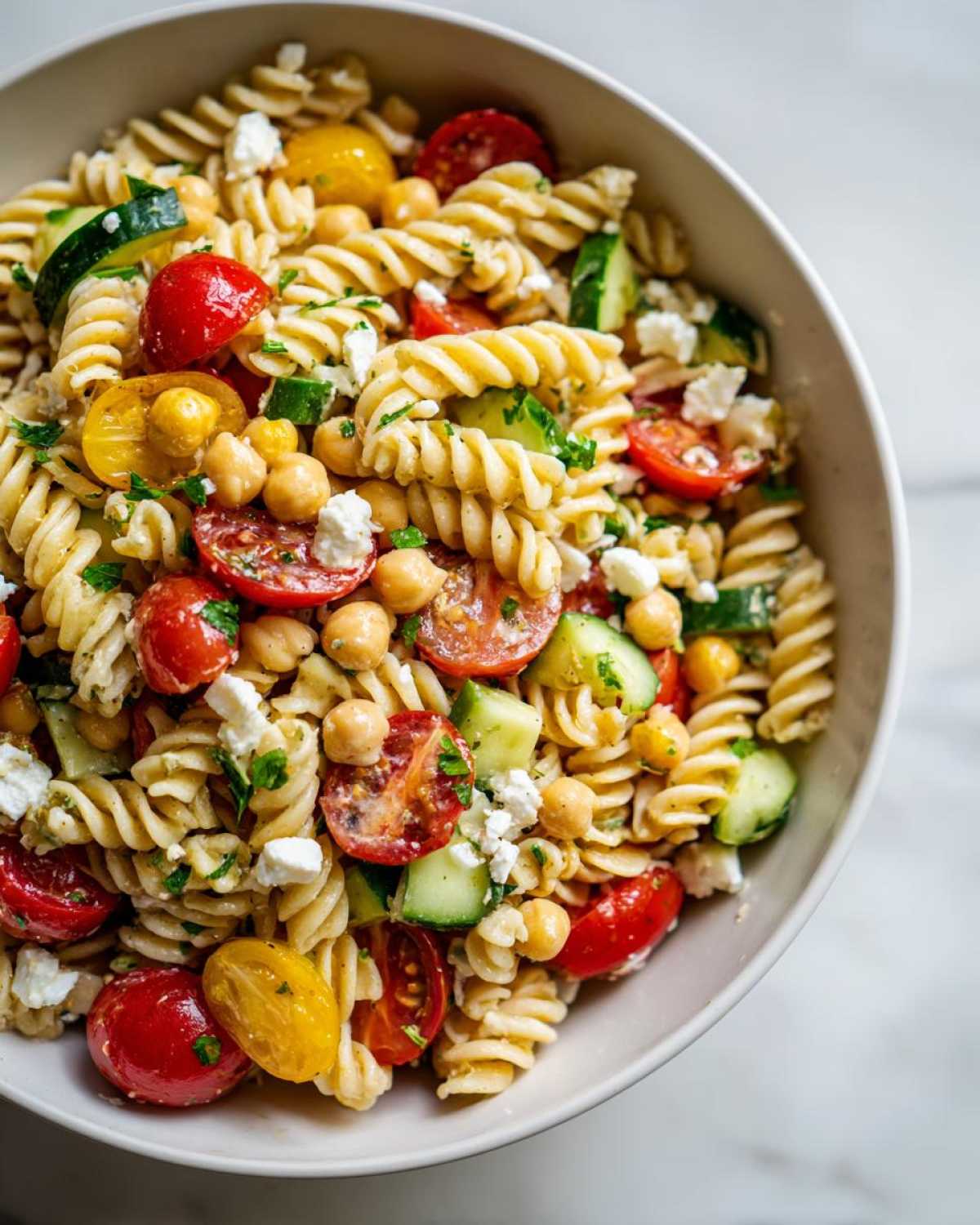 Close-up overhead shot of a bowl filled with vibrant Greek pasta salad with chickpeas, cherry tomatoes, cucumber, and feta cheese.