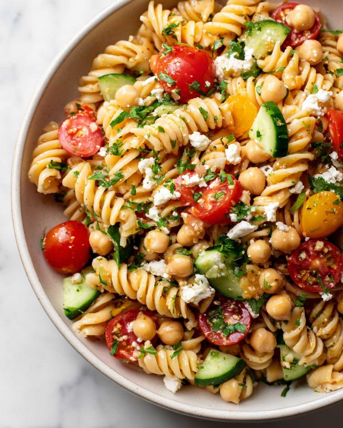 Close-up of a bowl filled with Greek pasta salad with chickpeas, cherry tomatoes, cucumber, and feta cheese.