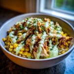A close-up of a healthy street corn chicken bowl topped with shredded chicken, corn, and a creamy green sauce.