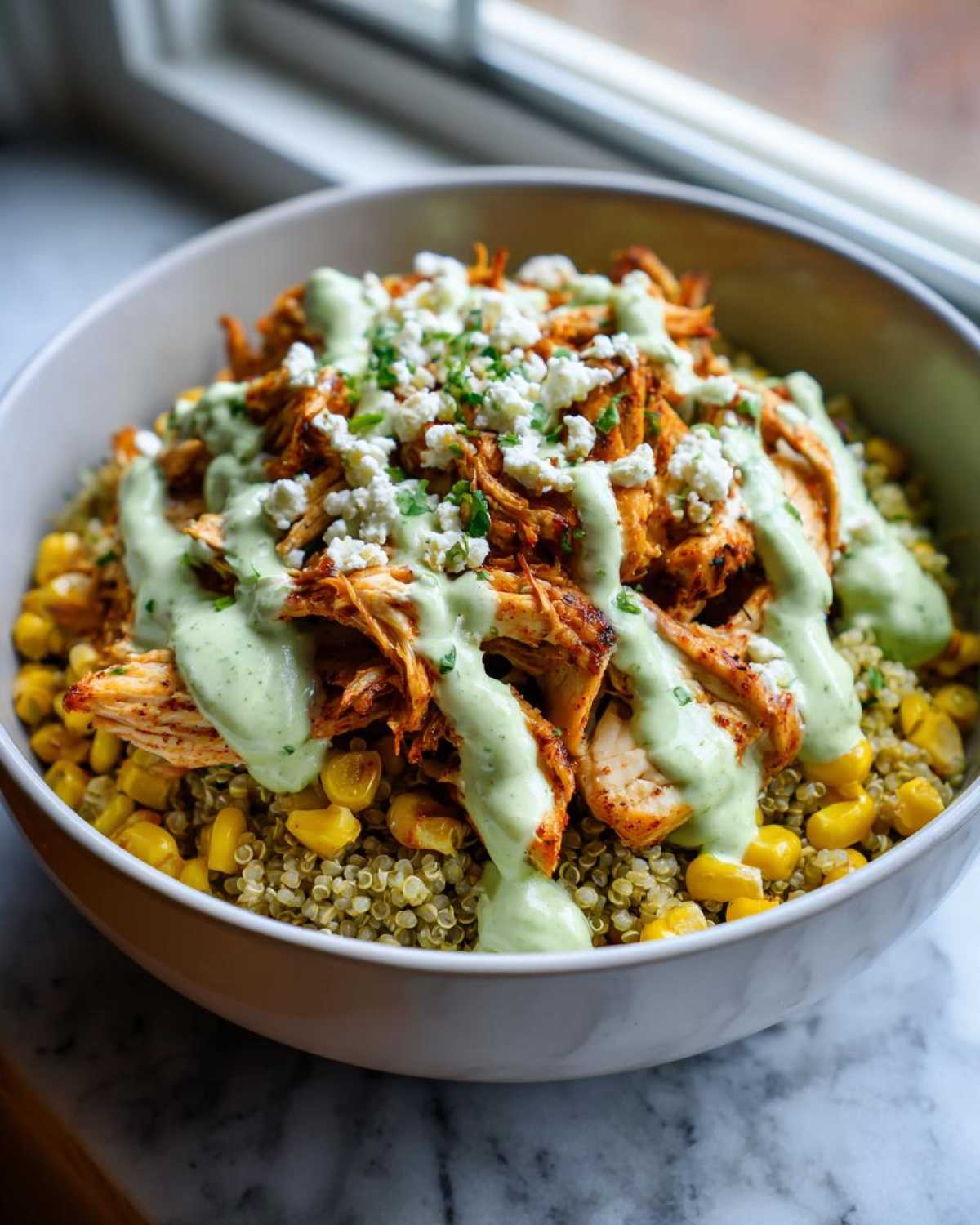 A close-up of a healthy street corn chicken bowl featuring quinoa, corn, shredded seasoned chicken, creamy green sauce, and cotija cheese.