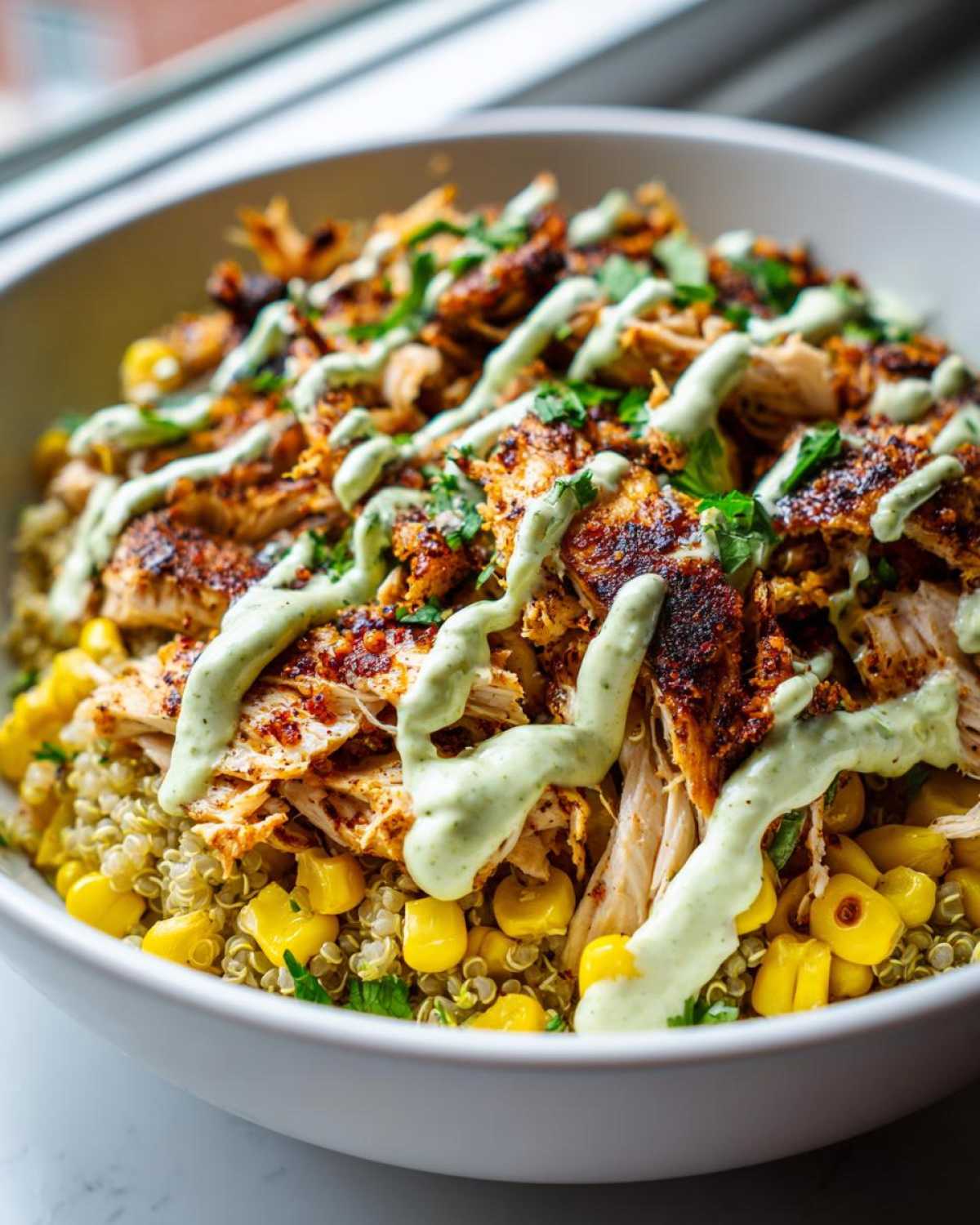 A close-up of a healthy street corn chicken bowl featuring seasoned chicken, corn, quinoa, and a drizzle of green cilantro-lime sauce.