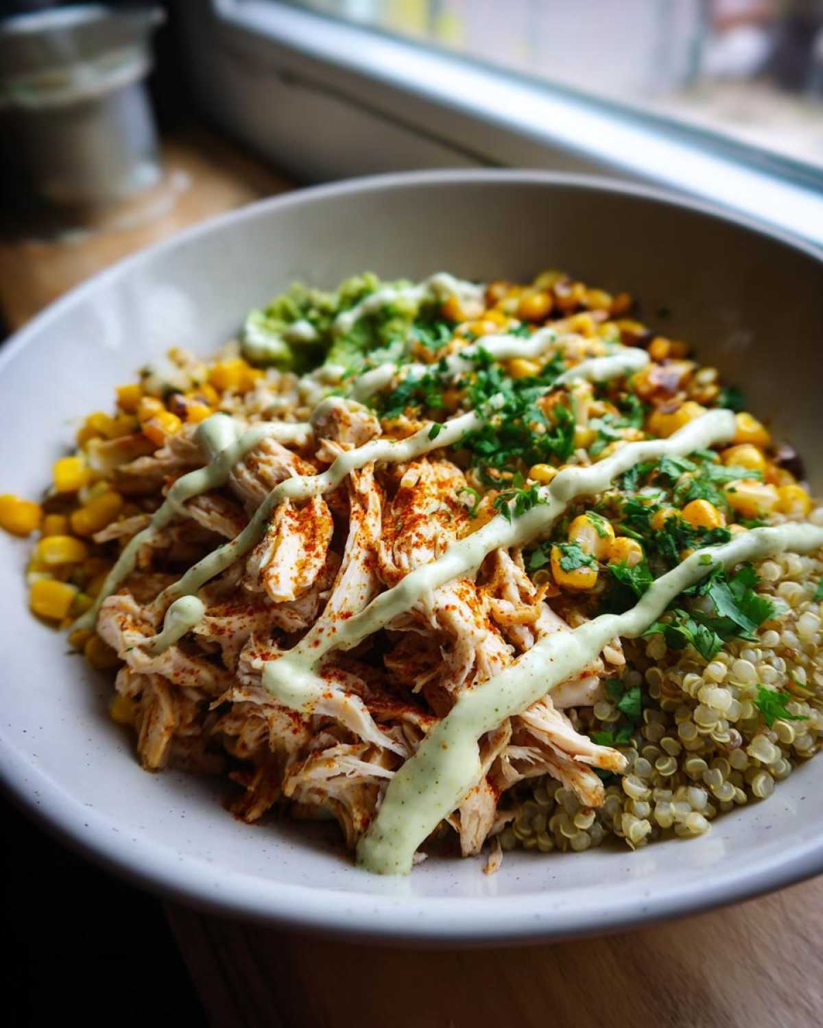A close-up of a healthy street corn chicken bowl featuring shredded chicken, corn, quinoa, and a drizzle of green sauce.