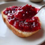 A spoonful of thick, bright red homemade strawberry jam being spread over a slice of white bread.