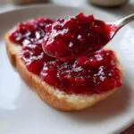 A spoonful of thick, bright red homemade strawberry jam being spread over a slice of white bread.