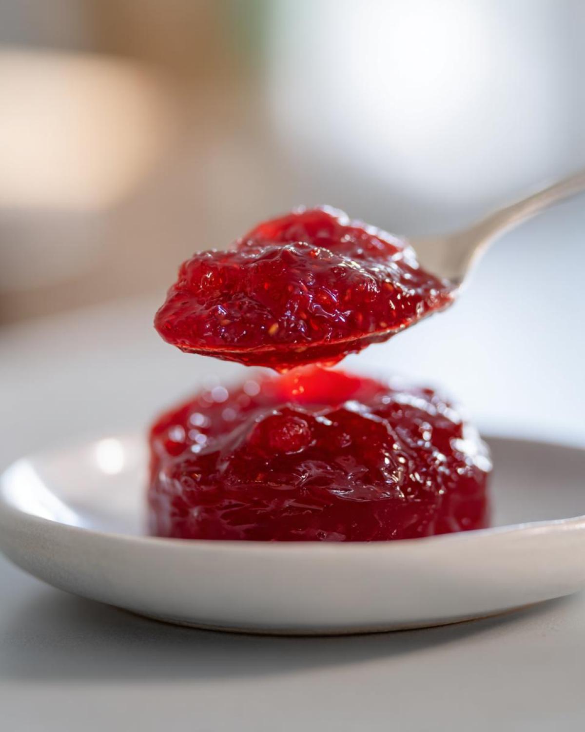Close-up of a spoonful of glistening Homemade strawberry jam being lifted above a small mound of the same jam on a white saucer.
