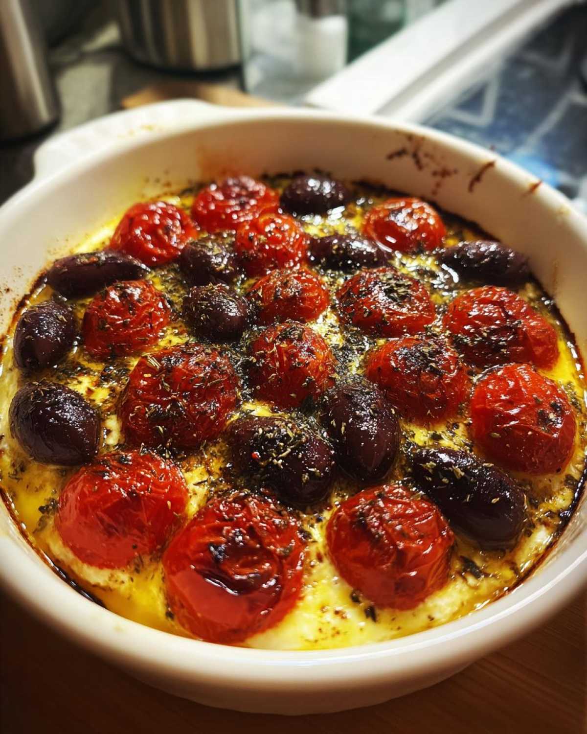 A close-up of hot Mediterranean baked feta dish bubbling in a white ceramic dish with roasted cherry tomatoes and Kalamata olives, seasoned with herbs.