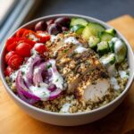 Close-up of a vibrant Mediterranean chicken bowl featuring sliced seasoned chicken breast over quinoa, topped with red onion, tomatoes, cucumber, olives, and white sauce.