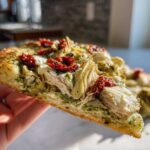 Close-up of a slice of Mediterranean chicken pizza topped with shredded chicken, artichoke hearts, sun-dried tomatoes, and pesto.