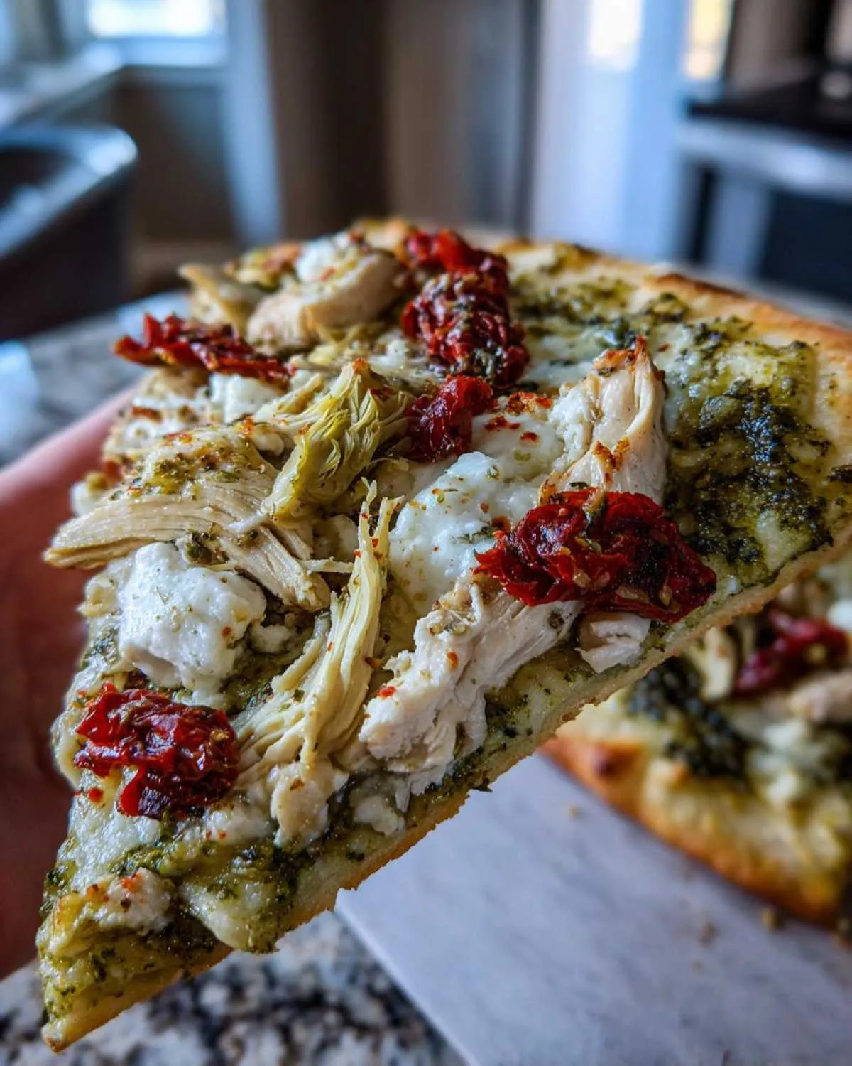Close-up of a slice of Mediterranean chicken pizza topped with pesto, shredded chicken, sun-dried tomatoes, and cheese.