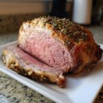 Close-up of a medium-rare roast leg of lamb, crusted with herbs, with one slice cut and resting beside it.
