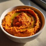 A close-up of rich, orange-red roasted red pepper hummus swirled in a white bowl, topped with olive oil.