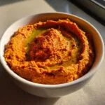 A close-up of rich, orange-red roasted red pepper hummus swirled in a white bowl, topped with olive oil.