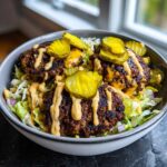 Close-up of a smash burger bowl featuring three charred patties over lettuce, drizzled with sauce and topped with bright yellow pickle slices.