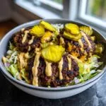 Close-up of a smash burger bowl featuring three charred patties over lettuce, drizzled with sauce and topped with bright yellow pickle slices.