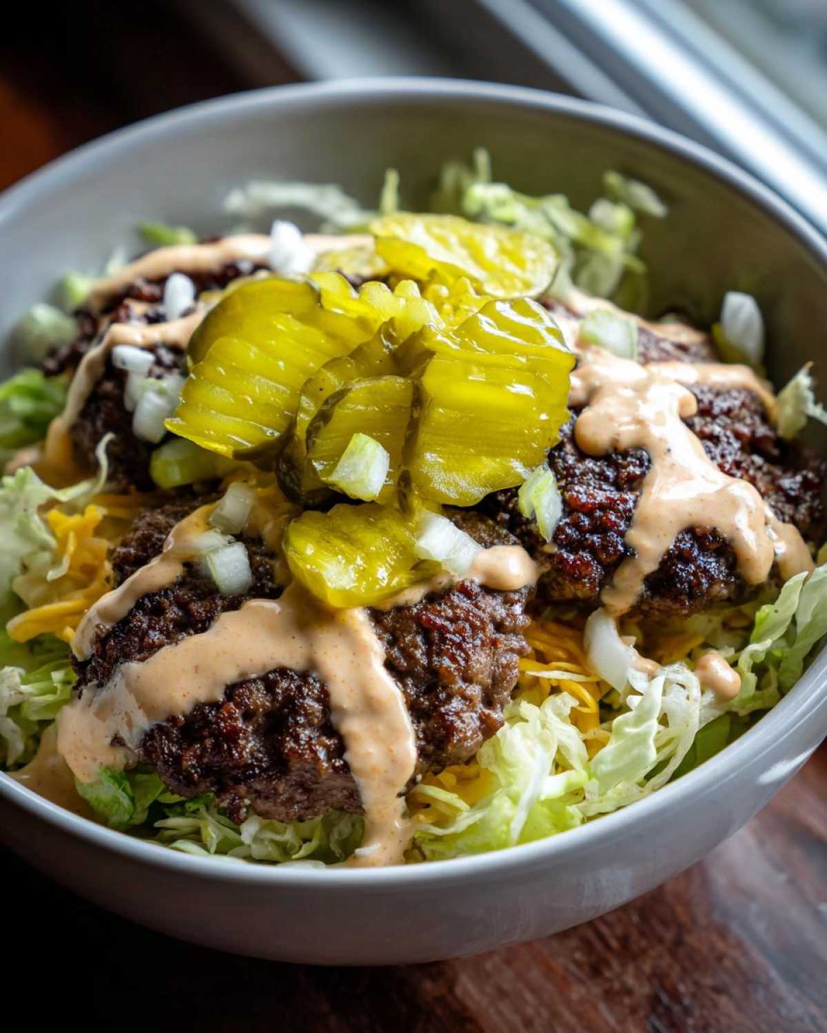 A close-up view of a smash burger bowl featuring seasoned ground beef patties, shredded lettuce, cheese, and a generous drizzle of sauce, topped with pickles.