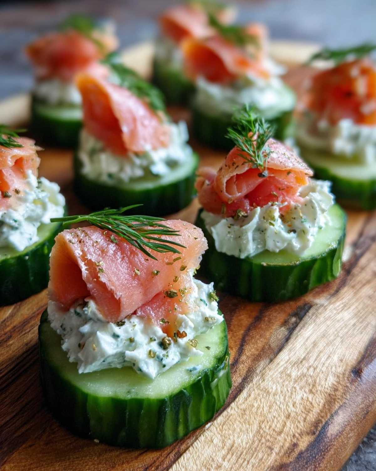 Close-up of several Smoked salmon cucumber bites topped with dill, arranged on a wooden serving board.