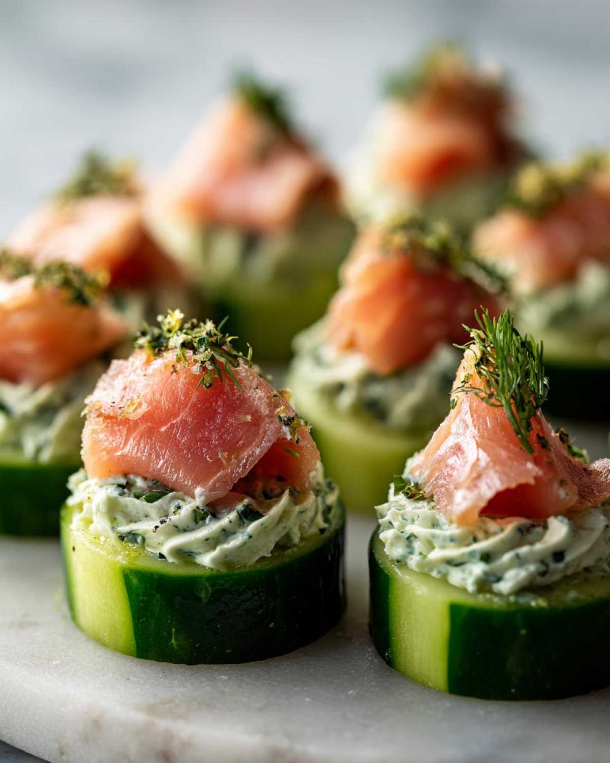 Close-up of several Smoked salmon cucumber bites topped with dill and cream cheese filling on a marble surface.