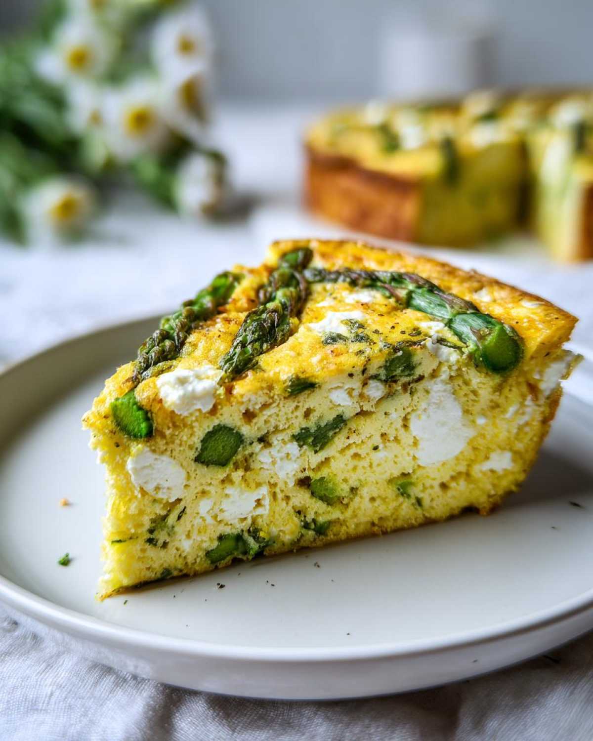 A thick slice of bright yellow Spring vegetable frittata featuring green asparagus pieces and white feta cheese, served on a white plate.