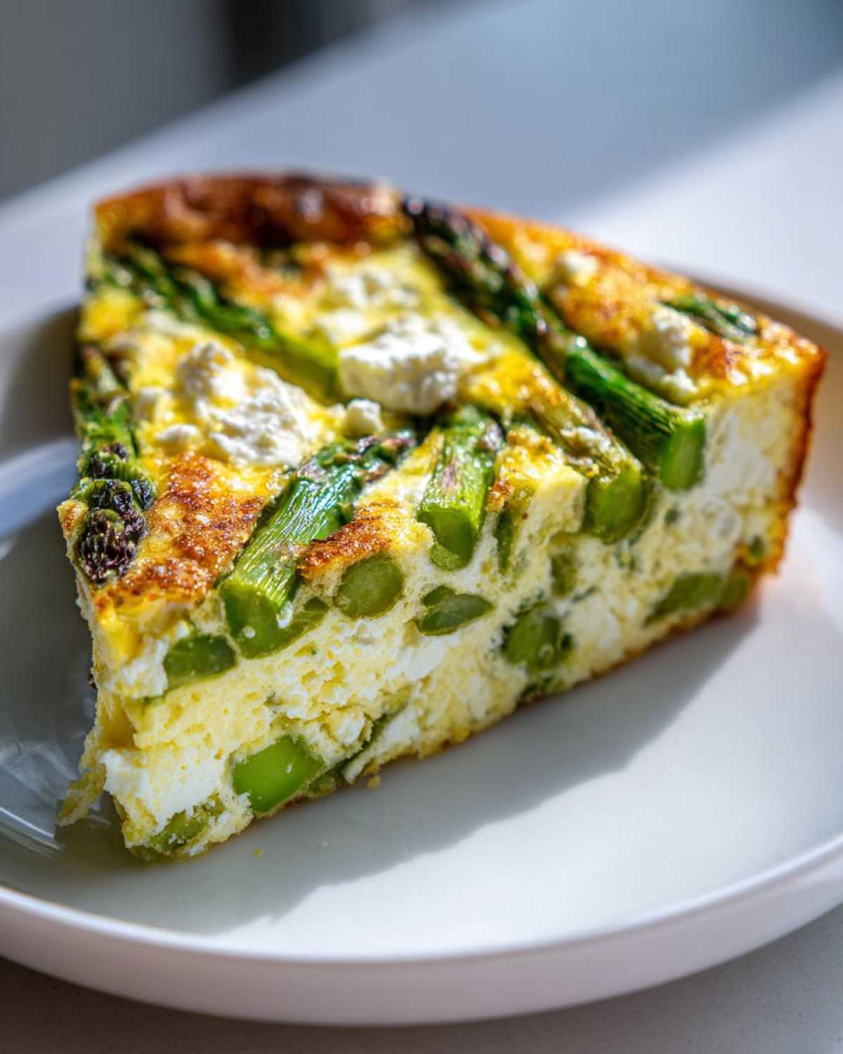 A thick slice of Spring vegetable frittata featuring bright green asparagus spears and chunks of white feta cheese.
