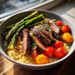 Close-up of a Steak couscous bowl featuring sliced medium-rare steak, pearl couscous, roasted tomatoes, and bright green asparagus.