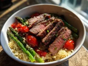 Close-up of a Steak couscous bowl featuring sliced medium-rare steak, roasted cherry tomatoes, and bright green asparagus spears.